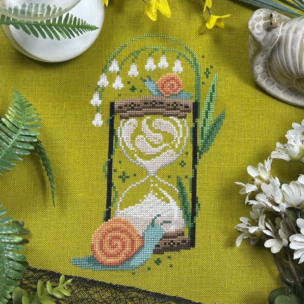 a cross stitch pattern of an hourglass with white lily of the valley flowers growing from the top while two green and peach snails sit in front and on top of the hourglass. The sand in the top part of the hourglass is shaped into ghosts. 
