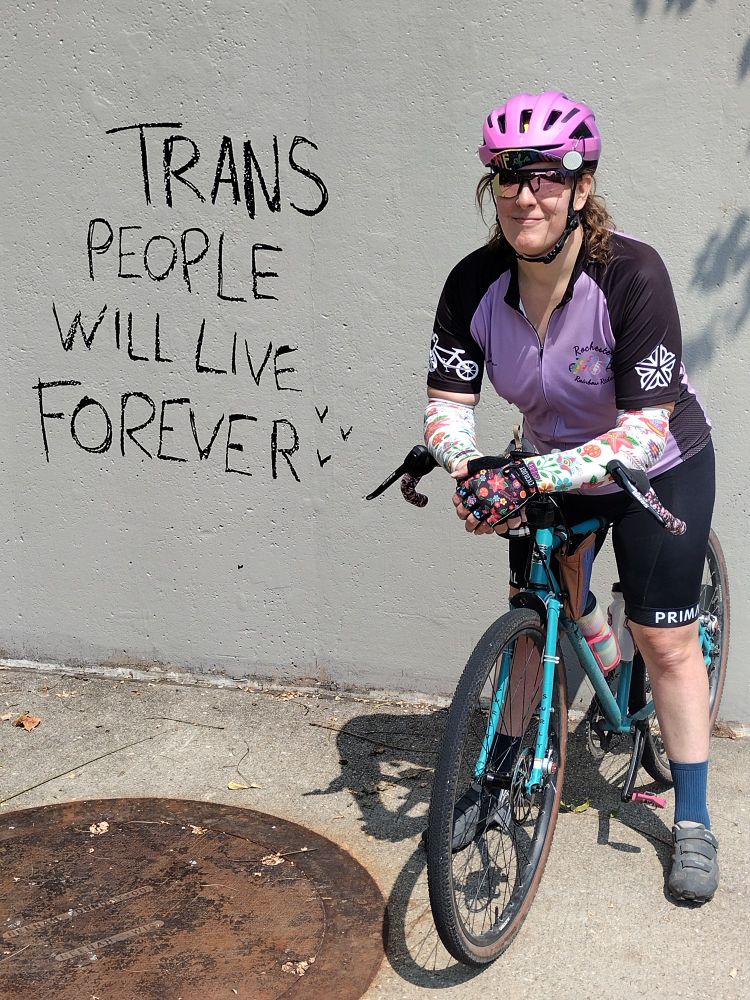 A cyclist sitting on her bicycle, resting her arms on the handlebars. She is next to a wall on which has been written "TRANS PEOPLE WILL LIVE FOREVER"