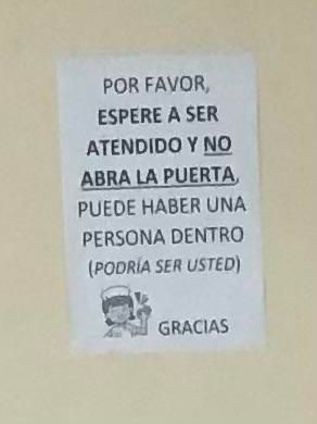 Cartel sencillo con un dibujo de una enfermera sonriente, pegado a una puerta, en la que se lee: por favor, espere a ser atendido y no abra la puerta. Puede haber una persona dentro (podría ser usted). Gracias. 