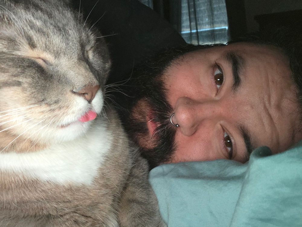 a selfie of me laying on my bed with my grey tabby cat Muse in the foreground, who has her eyes closed and her tongue sticking out just a little bit