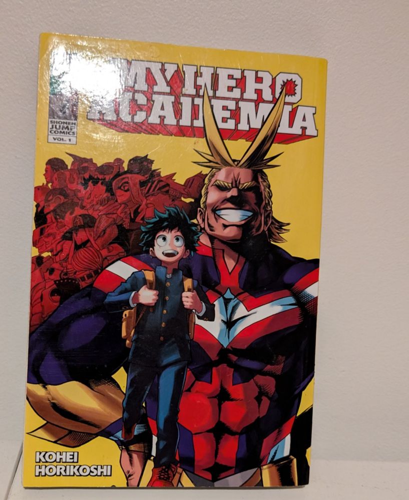 My hero academia vol 1 by Kohei Horikoshi. Midoriya a light skinned teenage boy with shaky dark green hair is smiling wearing his dark middle school uniform and red shoes, wearing a light brown back pack. Behind him smiling is All Might in buff form (light skin yellow hair miller with large bangs wearing his skintight red,white,blue, and yellow costume. Behind them are a number of proheroes in red Mt Lady, best jeanist, present mic, kamuy woods, backdraft, endeavor, 13, death arms, Nezu, ectoplasam, and a few nameless ones