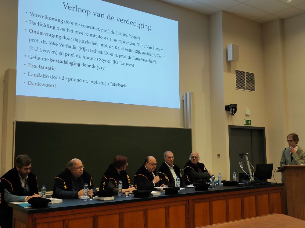 De foto toont de ondervraging tijdens mijn doctoraatsverdediging. Achter een lange tafel zitten zes professoren in toga. Een van hen, prof. dr. Karel Velle, heeft een microfoon vast: hij is een vraag aan het stellen. Ik sta rechts, achter een lezenaar, aandachtig luisterend.