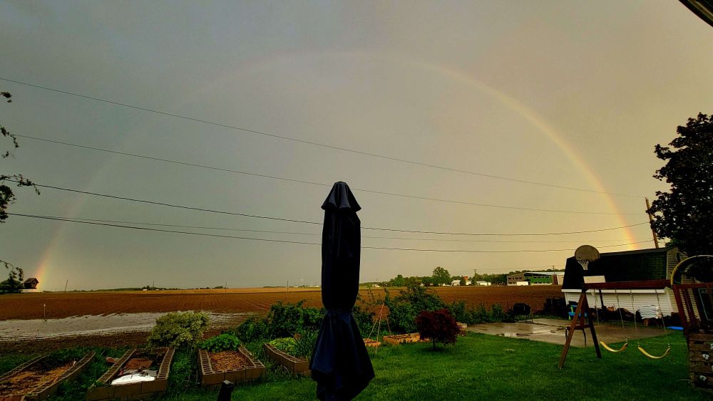A double rainbow over a farm field. A large table umbrella sticks up enthusiastically toward the middle of the middle of the rainbow. 