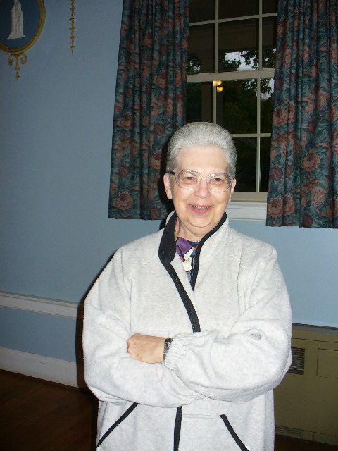 A smiling older women, in her late 70s, with short grey hair that has been slicked back. She is wearing a white zip up fleece and large clear rim aviator glasses. Her hands are lightly liver spotted. 