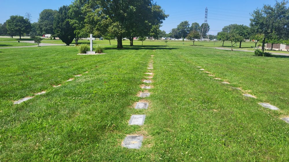 Rows of flat headstones are sinking into the grass. Some of them are almost completely covered. In the distance is a large white cross, about the size of a person and several trees. The grass is clearly mowed but no one is caring for the headstones. 