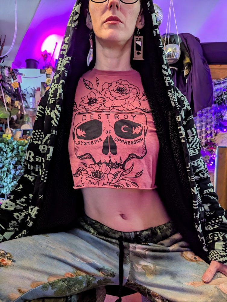 A woman fills most of the frame, posing with only below her eyes and above the knees in the frame. She's kneeling, wearing sweatpants and a fleece hooded cardigan with ASCII symbols scattered in neon green on it. She's also wearing a salmon-colored crop-top with an artistic skull and flowers, and the words "Destroy Systems of Oppression". She's wearing one guillotine earring and one sharp knife earring. 