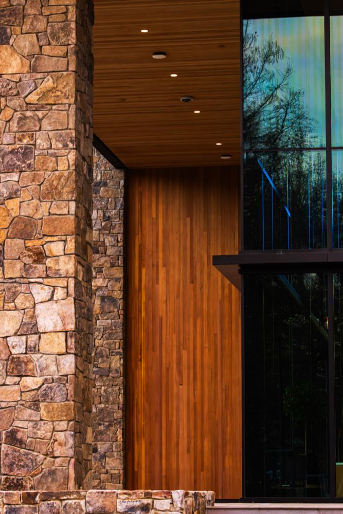 A modern architectural detail combines natural textures and materials. A tall, rough-hewn stone pillar occupies the left side of the frame, its warm brown tones varying in shade. To the right, a sleek wooden panel forms a doorway or facade element, its vertical slats creating a linear pattern. Behind the wood, large glass windows reflect a dark, wooded landscape with bare trees silhouetted against a dusky sky. Recessed lighting is visible on the wooden ceiling, adding a modern touch to the scene.