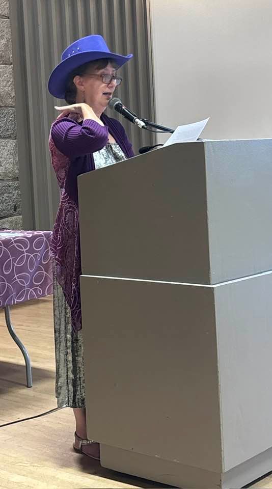 Janet Kvammen at the podium in Centennial Lodge reading into the mic and holding a page. Janet is decked our in a purple sweater and purple, sparkly cowboy hat.