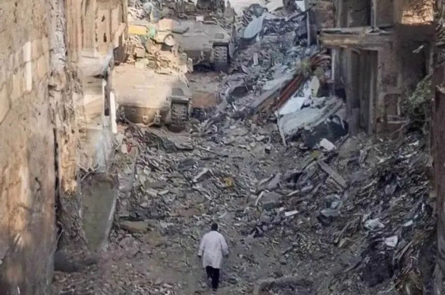 Un homme, en blouse blanche, marche sur les gravats, entre des bâtiments en ruine de la guerre. Il avance vers deux énormes chars qui lui bloquent la route. L’homme est le Dr Hossam Abou Safiya, directeur de l’hôpital Kamal Adwan, situé dans le nord de la bande de Gaza, attaqué par l’armée israélienne. L’image date de décembre 2024, le docteur a été arrêté ce jour-là.
