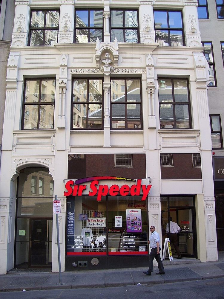 front facade of a white building with large windows and architectural flourishes. 

BIRTHPLACE OF FRANKLIN is across the top, referencing Founding Father Benjamin Franklin.

There is a Sir Speedy copycenter at the ground level, which is funny because he made his bones in his brother's print shop.