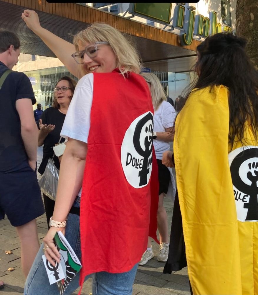 Een vrouw met een rode cape met het logo van de feministische actiegroep Dolle Mina staat lachend tussen andere mensen op straat. Ze heft haar vuist in de lucht in een strijdlustig gebaar. Naast haar staat iemand met een gele cape met hetzelfde logo. Op de achtergrond zijn meerdere mensen in gesprek en een winkelgevel zichtbaar.