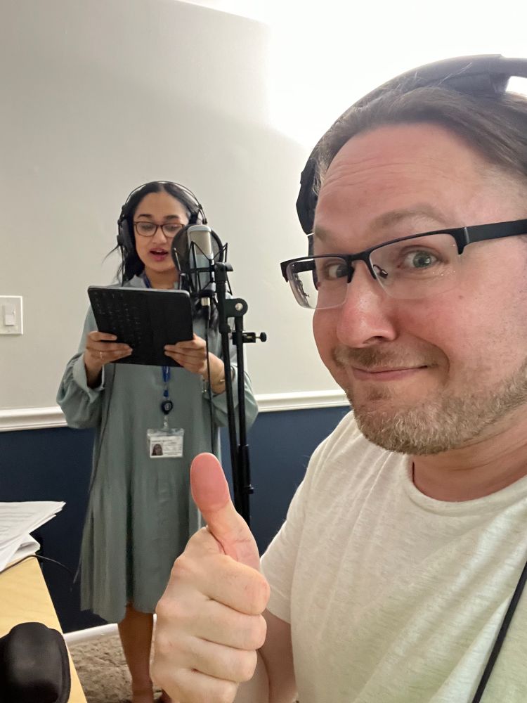 I am in the foreground smiling and giving a thumbs up for a selfie while one of they young ladies is in the background holding sheet music and in the midst of singing  into a microphone
