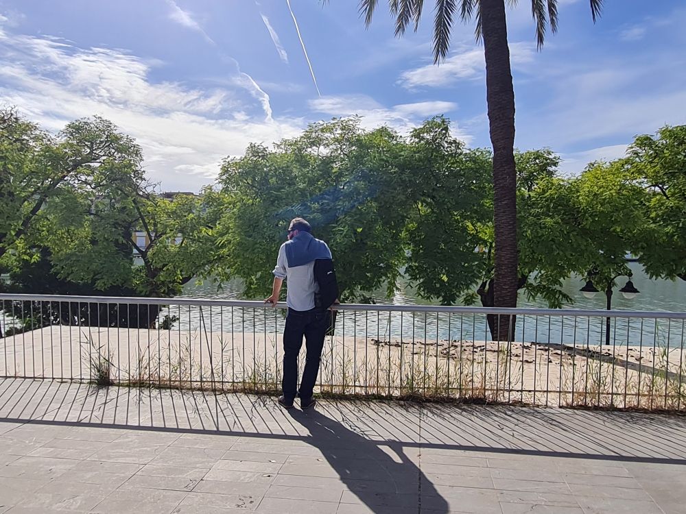 My husband Jamea leaning over a railing and looking for fish in the river. There are green trees along the bank and blue sunny skies above