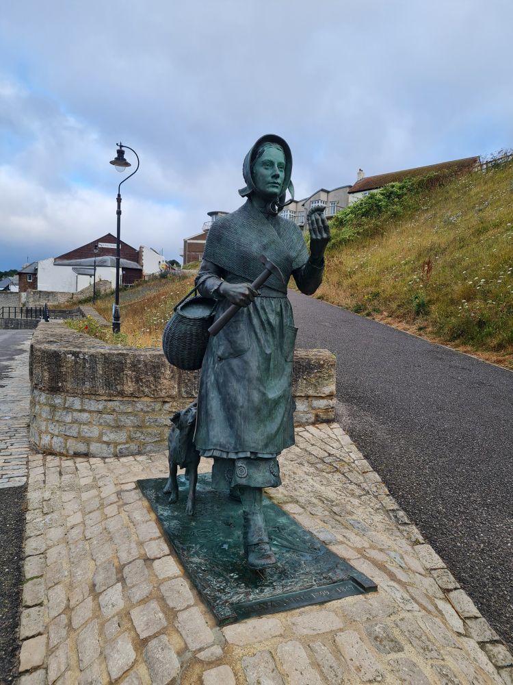 Statue of Mary Anning at Lyme Regis striding towards the beach with hills and paths behind her