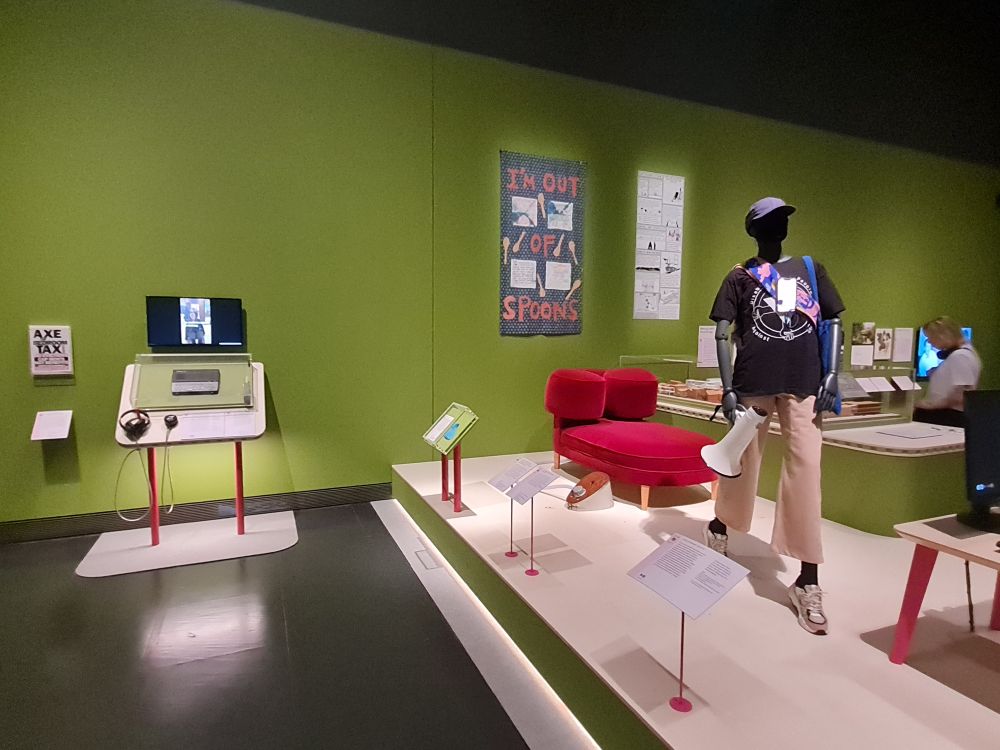 Image of the exhibition Design and Disability at the V&A showing a mannequin holding a loudspeaker and wearing a dark blue t-shirt, pale trousers and a blue cap. A phone is hanging from its neck showing how disabled people can protest through a proxy. Behind the figure is a red hug chair designed to provide sensory comfort.  On the wall is an artwork showing spoons and reads "I'm out of spoons". On the left is a video with a keyboard displayed underneath. 