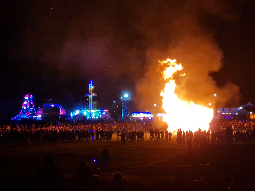 Multicoloured lit up fairground rides with a large bonfire on the right surrounded by hundreds of people staring at the fire
