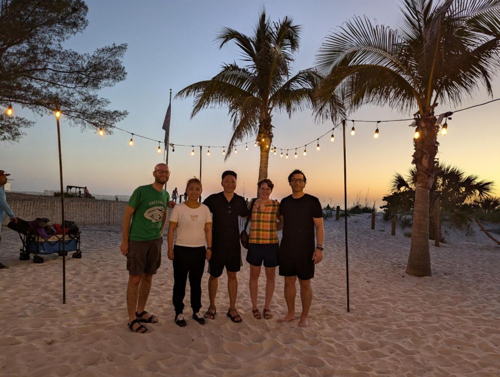 Five people stand together, posing for the camera, on a beach with palm trees and lights.  The sunset and hanging lights cast a warm glow.