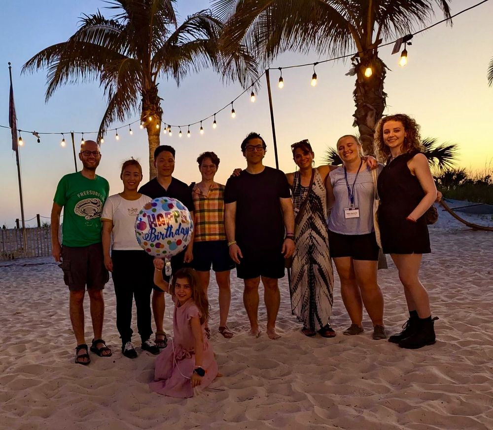 Eight people stand together on a beach, posing for the camera, while a ninth person sits in front of the row of people holding a "Happy birthday!" balloon.  Two palm trees and a sunset complete the beach background.
