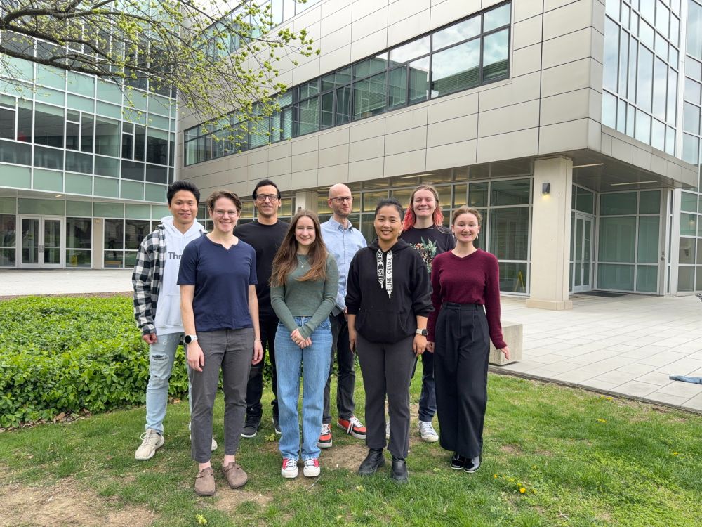 Eight people stand in two even rows in front of a glass and concrete building.  A tree stretches its branches in the top left corner, and greenery abounds on the ground.