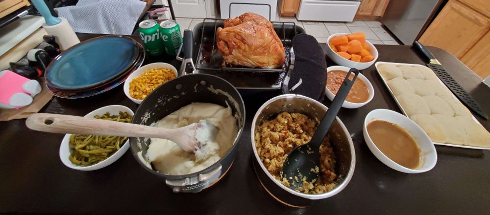 A counter filled with a Turkey Day spread: stacks of plates; 2 cans of sprite; corn; green beans; mashed potatoes; stuffing; yams; baked beans; gravy; bread rolls; and of course, a turkey