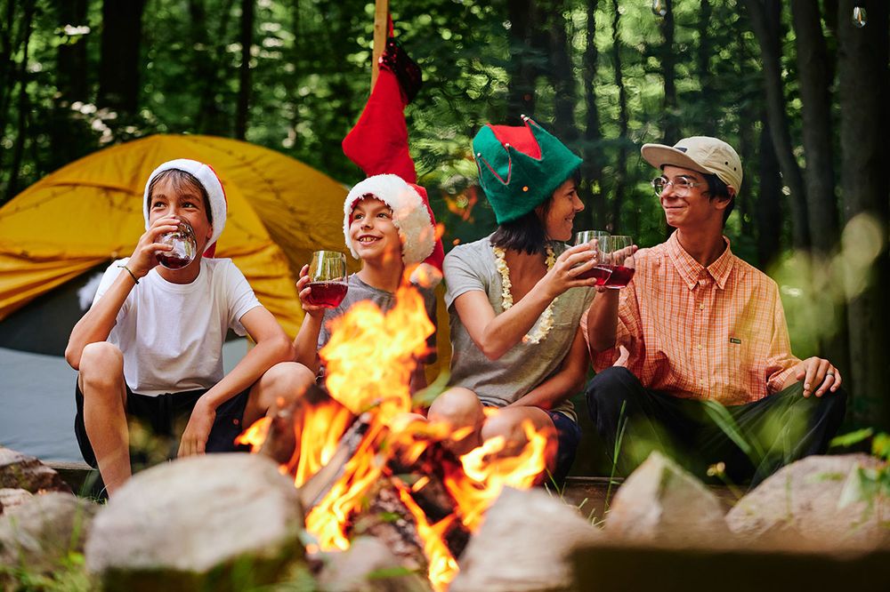 Four persons in front of a fire. Two of them are toasting. Three are wearing holiday hats. There is a tent in the background. It’s summer.