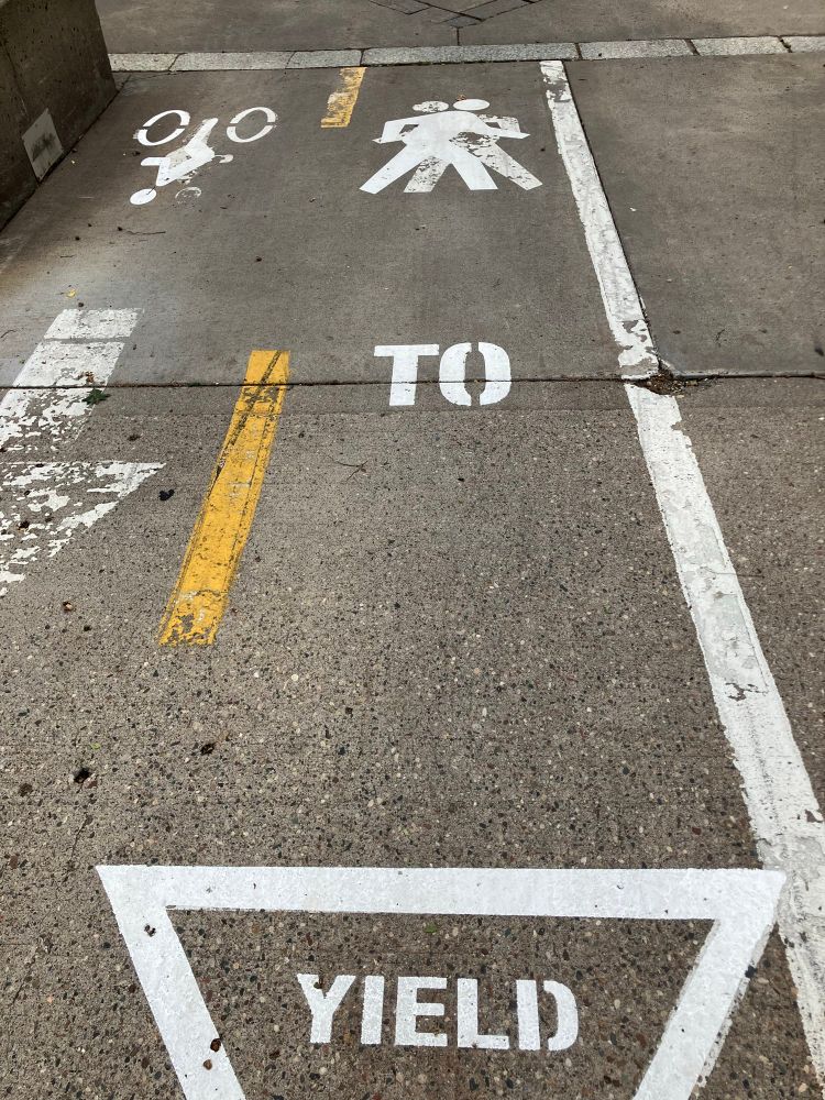 Stencil in pedestrian lane: in the foreground, YIELD sign; farther away, a worn-down pedestrian symbol facing left, with a fresh layer superimposed but facing right. They look like partner dancers.