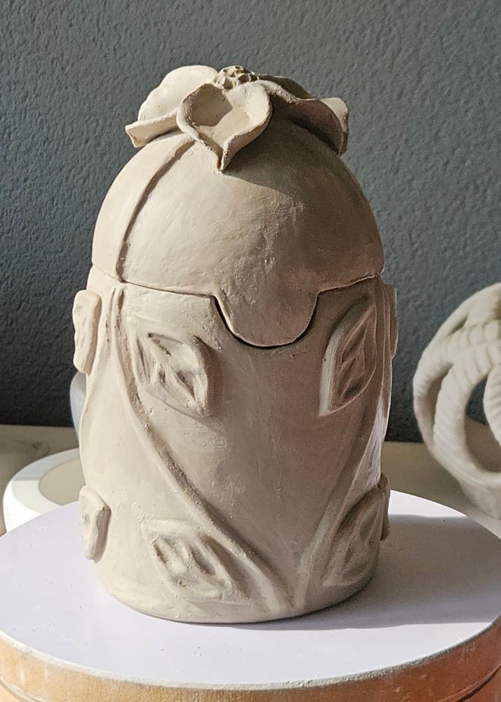 Clay vessel with sculptured flower on top and leaves and branches on the side. 