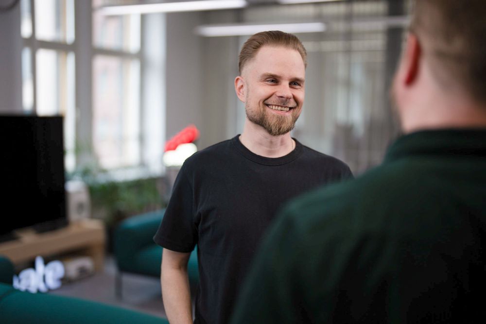 A smiling man in a black t-shirt stands in a modern office, speaking with a colleague. The atmosphere is relaxed and friendly, with soft lighting and a blurred background showing sofas, plants, and large windows.