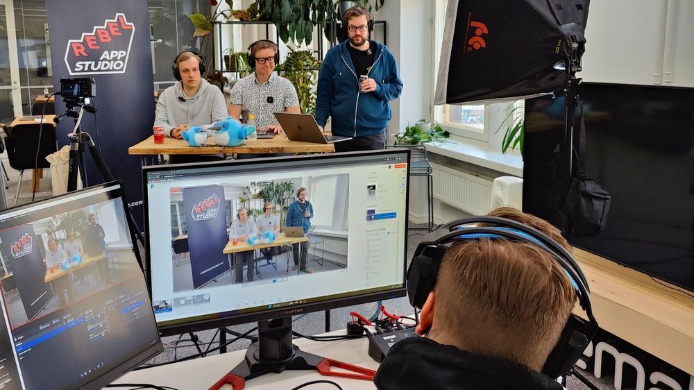 Behind-the-scenes of an accessibility meetup at Rebel App Studio office. Three speakers sit at a table with a Rebel banner behind them, while a producer manages the live stream in the foreground. A camera and monitor display the broadcast, with plush mascots and drinks on the table.