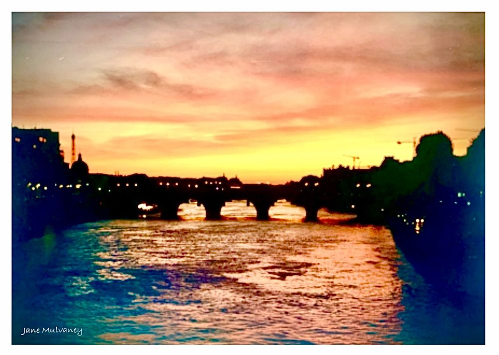A photo of a sunset over the Seine, Paris. A bridge & surrounding buildings are silhouetted. The orange & yellow sky tones are reflected on the choppy surface of the river. The lower corners of the old analogue photo are fading to blue & purple, and bleeding into the river surface.