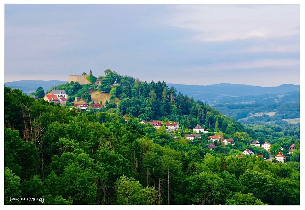 A view of Burg Lindenfels in Germany, with an old castle on top of the hill, houses on the slopes, and surrounded by forest. There are more hills and forest in the distance. The sky is partly cloudy.