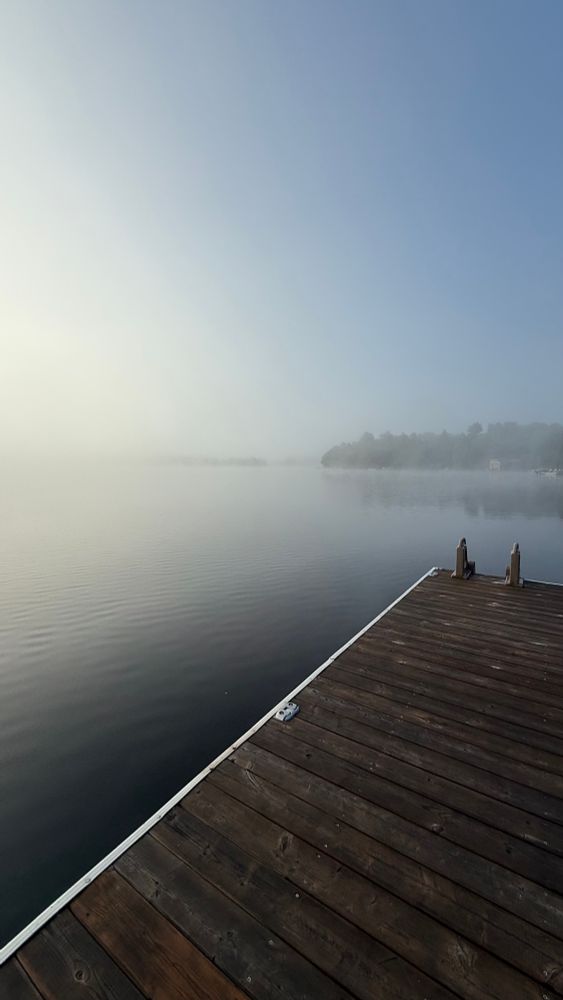 A calm lake in Ontario Canada with some of a dock in the foreground and morning mist on the lake. 