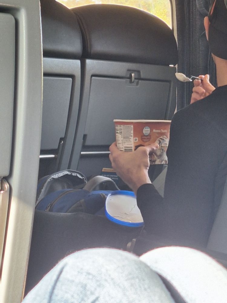 Guy in an aisle seat on the train, in conversation with the person in the windows seat, eating a spoonful of ice cream out of a 1.5 qt container of ALDI's brand moose tracks ice cream