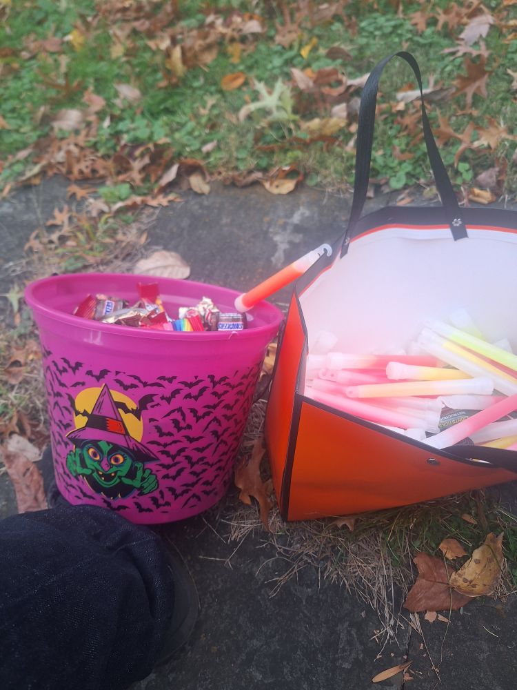 Pink bucket full of mini candies, with a cartoon witch and bats on the front, beside an orange bag full of glowsticks. 