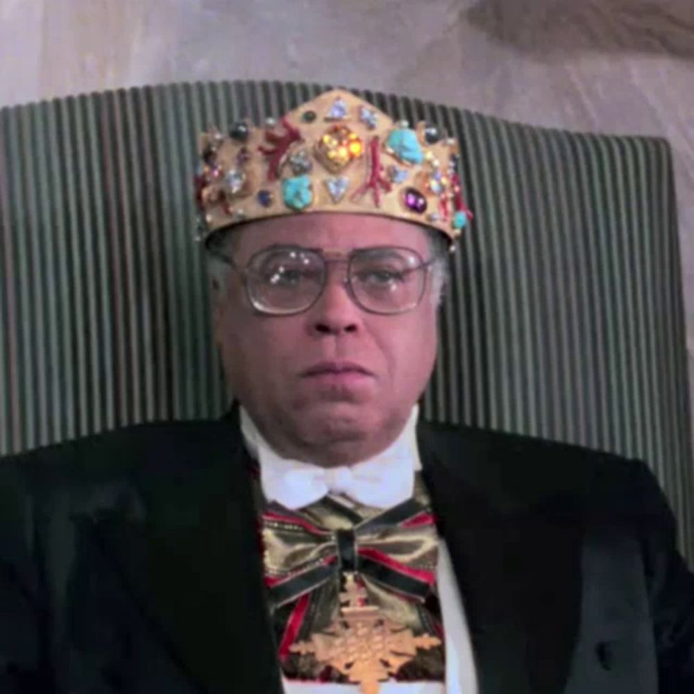 a man wearing a crown and a bow tie is sitting in a chair