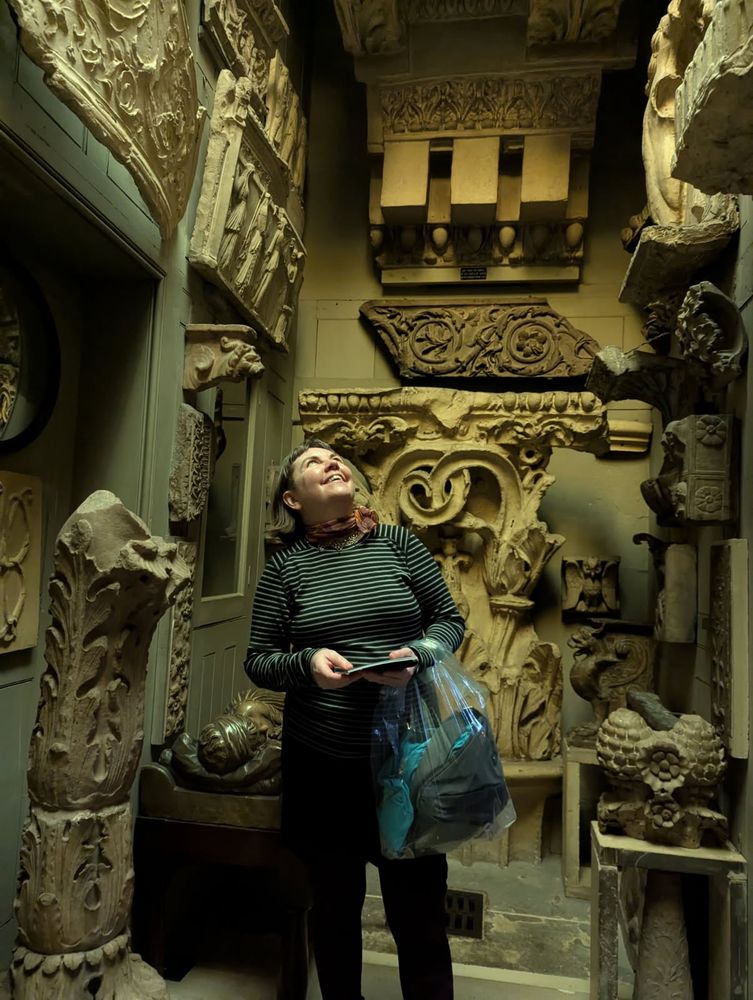 Kristin is wearing a black and grey striped shirt, standing in the center of a narrow space, looking up. The walls up and around her are covered with pieces of statuary from around the world.
