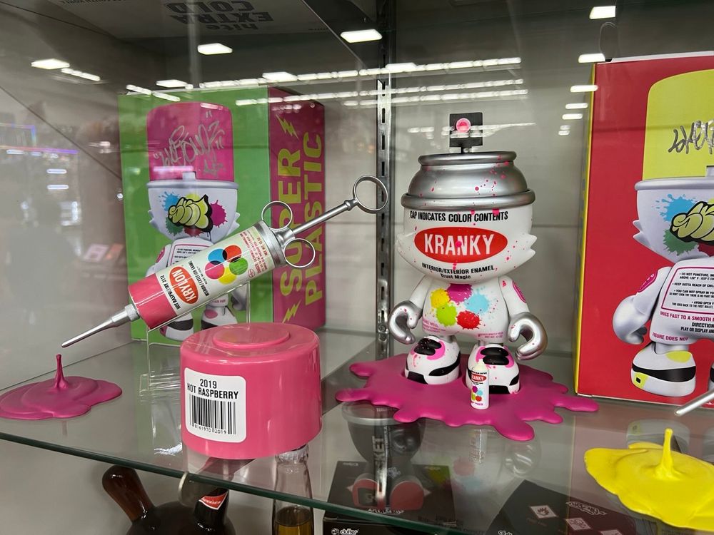 A display case featuring Superplastic designer toys is shown. One figure, labeled "KRANKY," resembles a paint can with a splash of colorful paint on its body and base. Next to it is a syringe-like paint marker with "HYPE COLOR" branding and colorful paint splashes. The packaging in the background is bright green and pink with graffiti-style artwork.