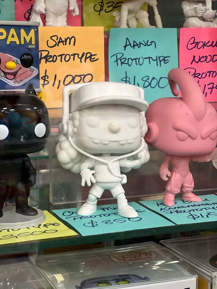 A display shelf shows several collectible prototypes, including a white figure with a hat and headphones priced at $850. Other prototypes include a black batman  figurine on the left, a pink figure on the right, and additional figures in the background, each with handwritten price tags such as "Aang Prototype" for $1,800 and "Goku Prototype" for $1,700. Brightly colored notes with the names and prices of the figures are placed behind each one