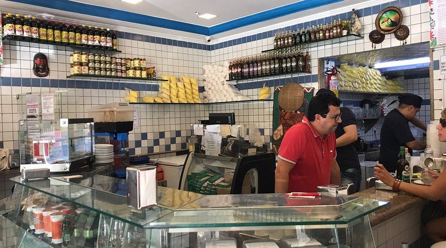 foto do restaurante citado. a foto mostra o balcão e duas das paredes, todas cheias de prateleiras com produtos do norte. dentro do balcão, uma pessoa atende um cliente sentado num banco e atrás de tudo duas pessoas preparam coisas na cozinha