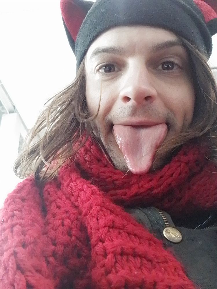 Miu is in a black cap that has cat ears stitched onto it and red handmade scarf and coveralls stick out their tongue. Selfie is in the library