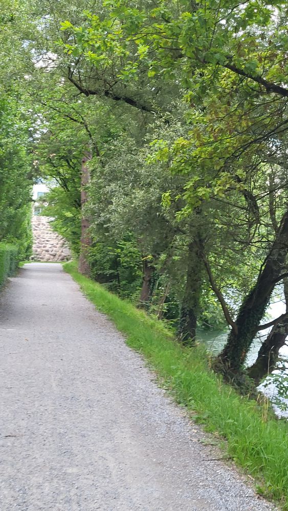 Gravel walking path lined by trees that part to show a foundation built of very large rocks der in mortar. The edge of a river is visible between trees on the right. 