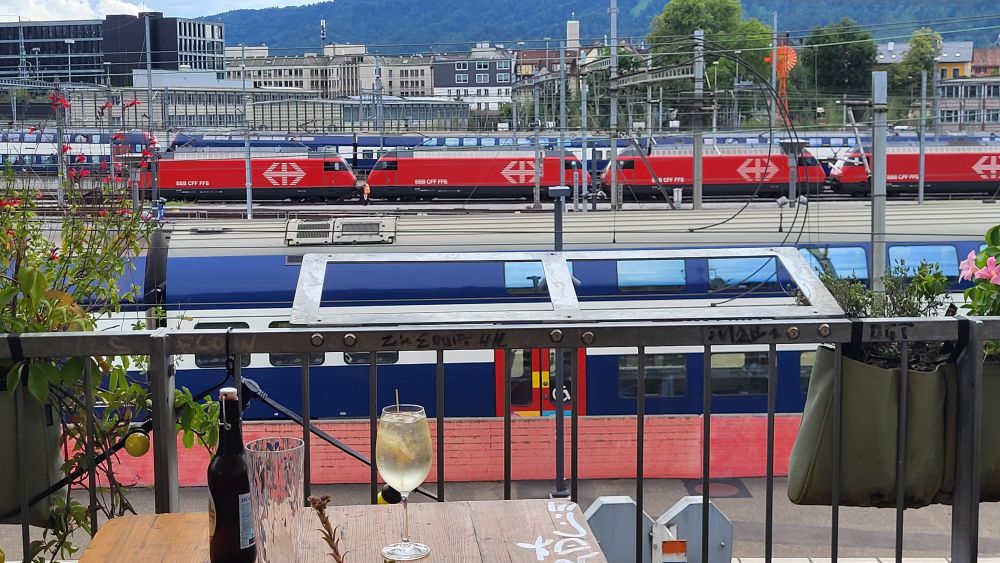 In the foreground, a table with a beer bottle and a wine glass next to a railing. In the background, several red and white and blue trains. Beyond that, tallish buildings.