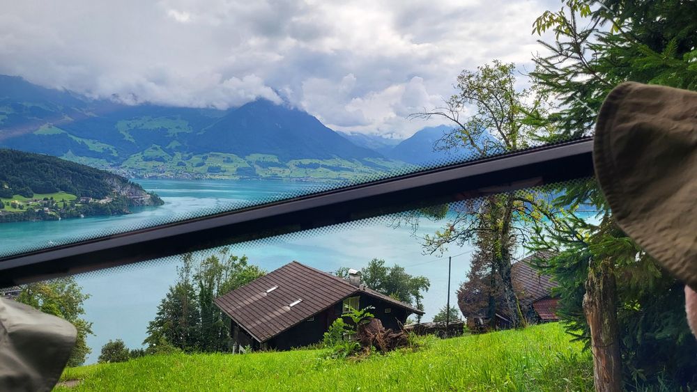 Photo of a green meadow with a farmhouse and an aquamarine lake and mountains in the distance through a train window. The window is broken by a frame that is at a very steep angle, contrasting with the horizontal of the lake shore.