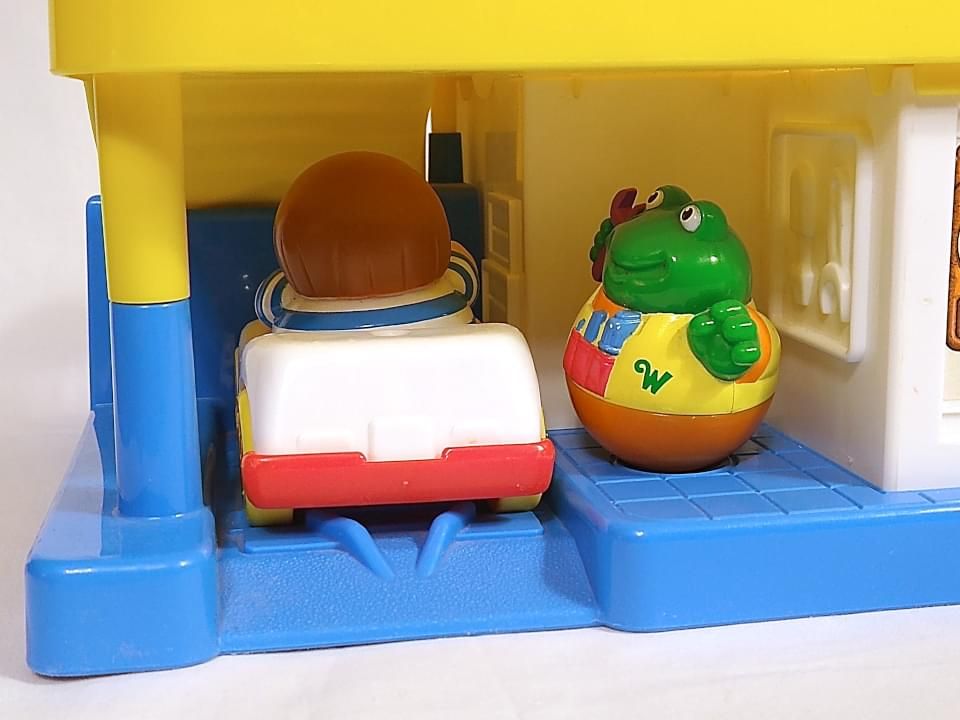 Mechanic's bay of Weebles Gas Station playset. Highway (boy with brown hair in a pageboy cut and no legs wearing a blue and white striped shirt) sits in a one-seat white car with a red underbody parked on a ramp. Uggie Wobbleworks (green frog with no legs wearing an orange coverall and a yellow vest with a red pocket containing blue tools and holding a purple wrench) sits next to a tool rack facing the car.