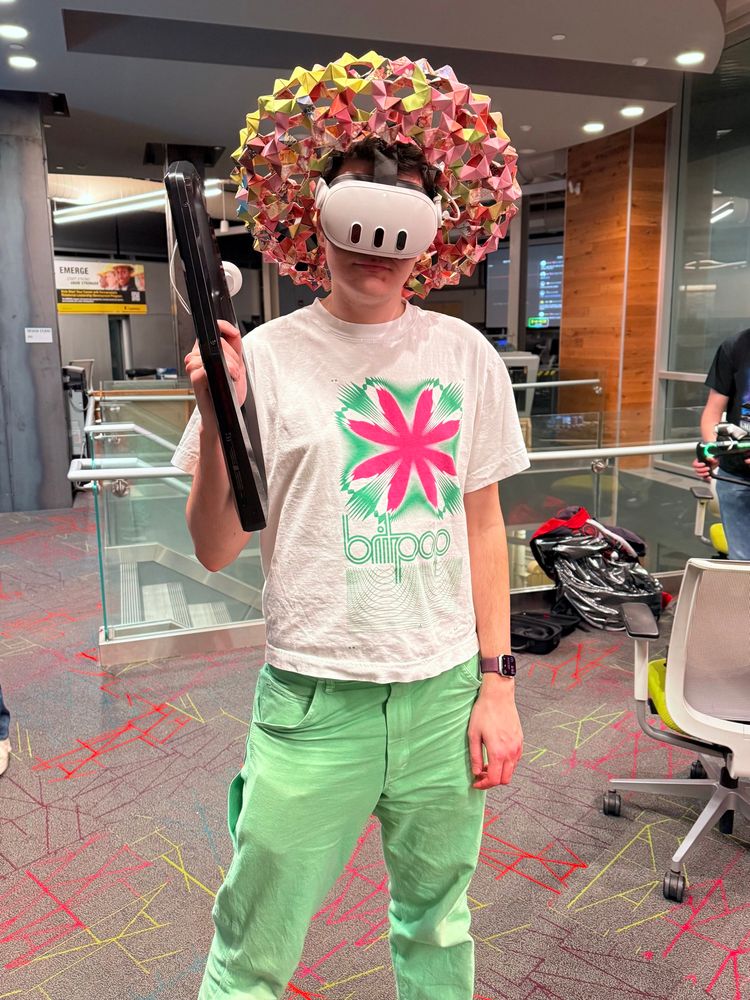 Me wearing green pants, an A. G. Cook Britpop shirt, a pink-orange origami wreath(?), and a VR headset, and holding a plastic toy rifle-looking thing