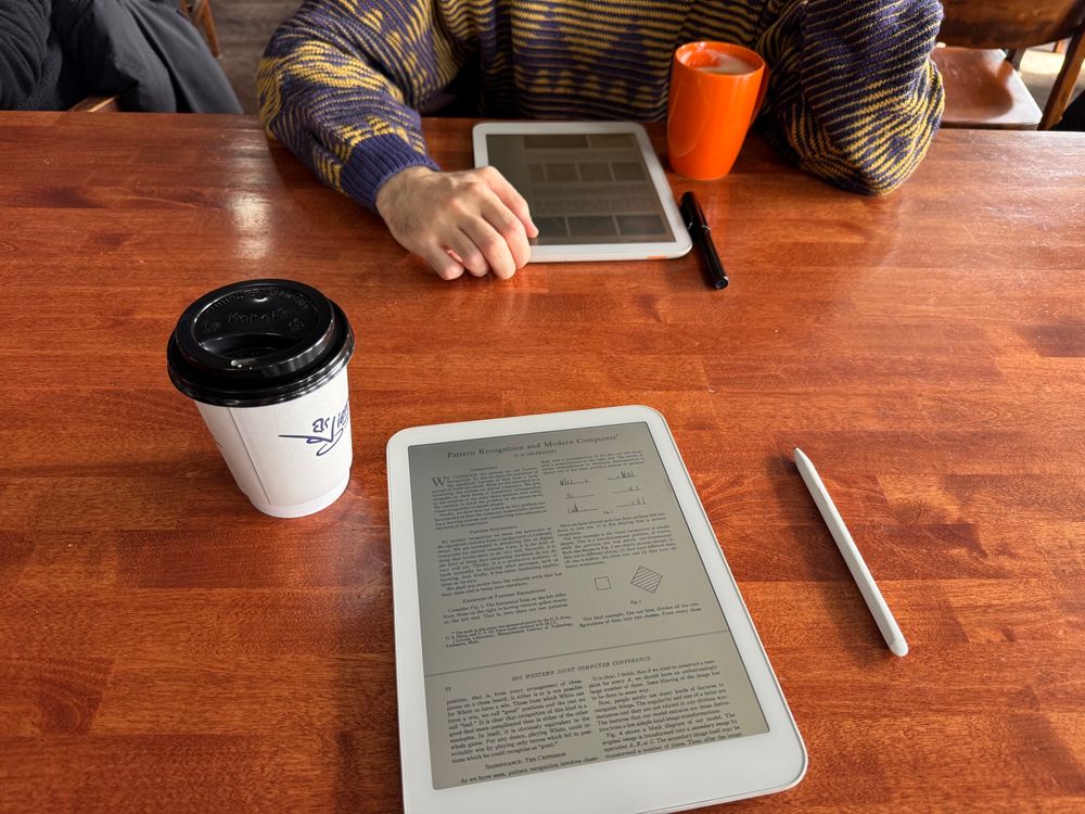 A wooden tabletop at a coffee shop with two coffees, POV, a daylight tablet in front of me and the person sitting across from me, whose face is not visible