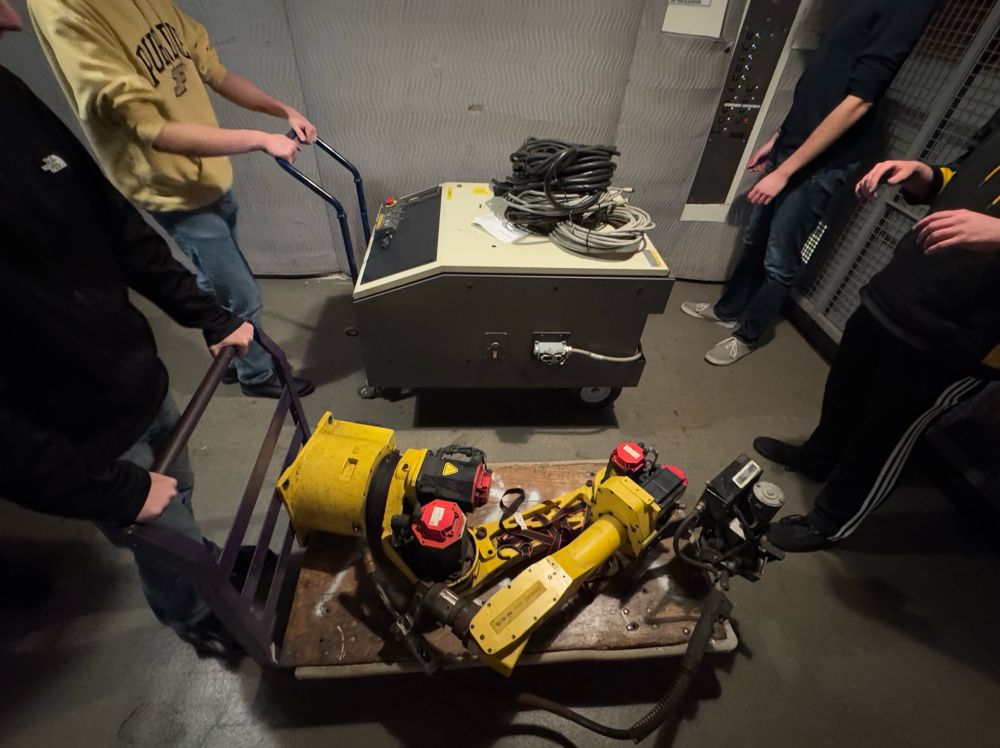 4 people carrying a yellow robot arm and a large white control board on two dollies