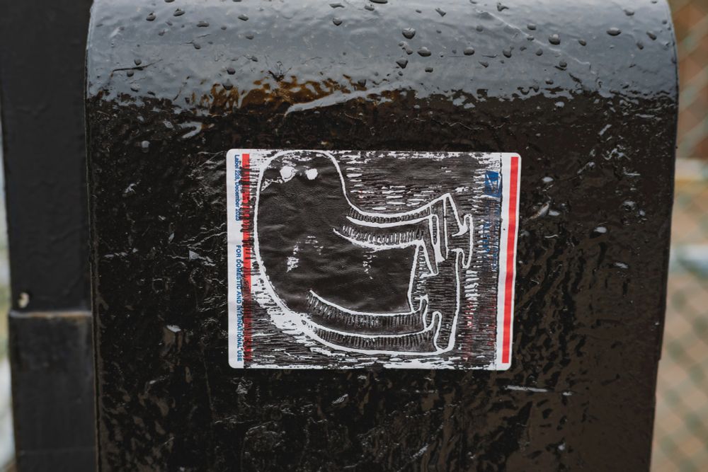 A stamp of a ghost all in black ink on a USPS shipping label stuck onto the side of a traffic control box.
