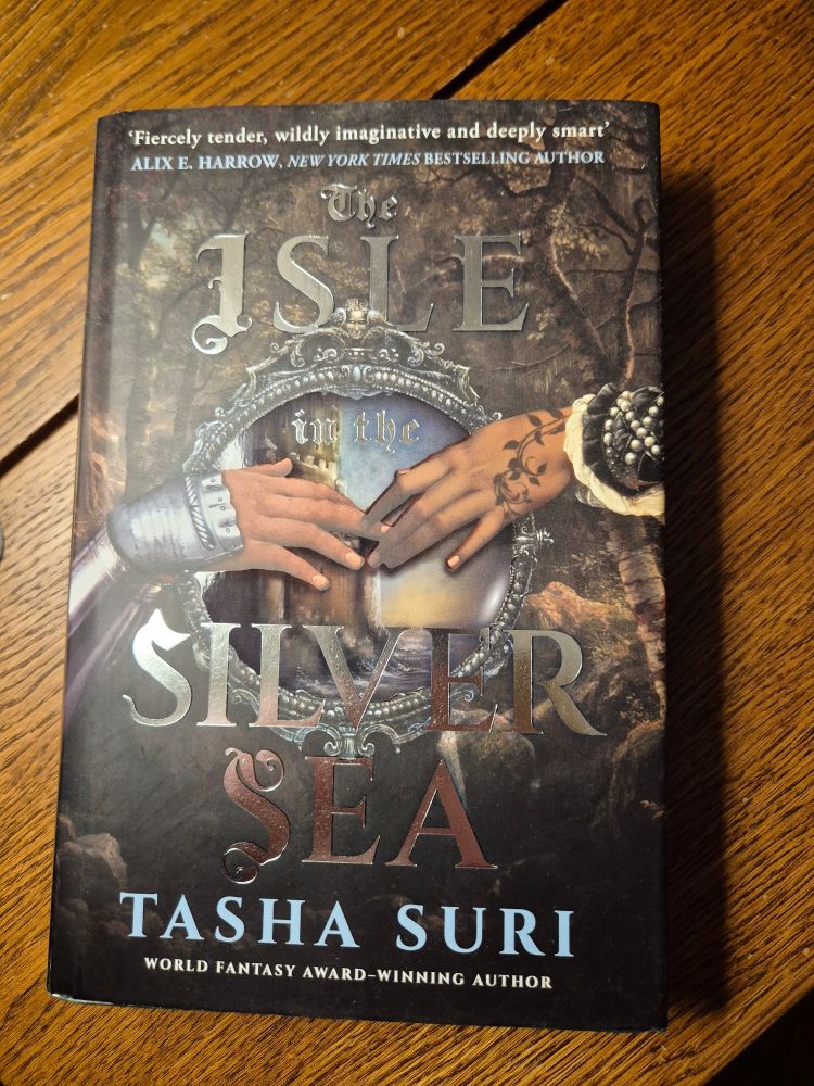 Physical copy of the book. The Background shows woods and in the center is a mirror? that shows a castle by the sea. In the foreground are two hands reaching for each other. The one on the left wears armor, the one on the right wears cloth adorned with pearls and is tattooed with curling vines.