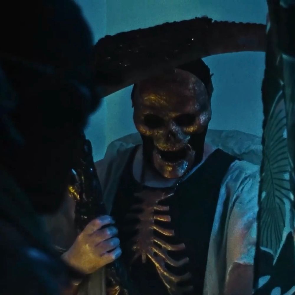 a still from the movie Ribspreader (2022), a man with a golden skull mask and large scythe is wearing a large necklace shaped like a ribcage that covers his whole chest